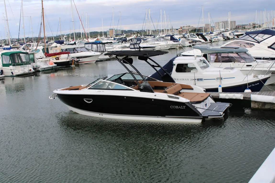 Cobalt boat docked at a marina surrounded by various yachts and boats under a cloudy sky, reflecting on the water.